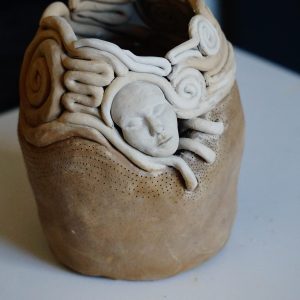 A handmade coil pot with a solid, brown bottom that turns into swirly coils outlining a woman's face.